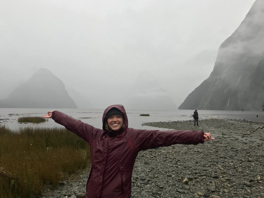 a photo of a woman holding out her arms and smiling, with misty mountains and water in the background